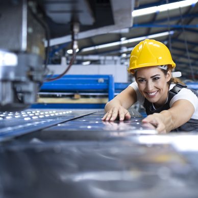 Female factory worker in protective uniform and hardhat operating industrial machine at production line.