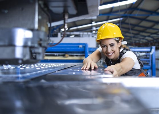 Female factory worker in protective uniform and hardhat operating industrial machine at production line.